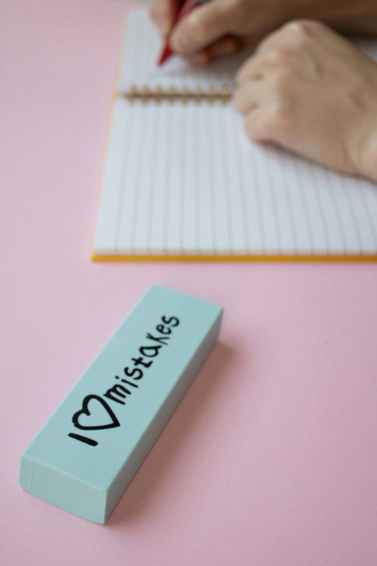 Close-up of hands writing in a spiral notebook with an eraser on a pink surface.